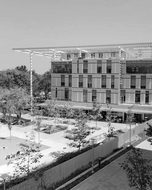 A modern multi-story building with a flat roof and extensive glass windows, surrounded by a landscaped outdoor area featuring young trees, paved walkways, and neatly maintained garden beds. The garden