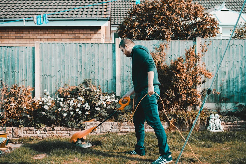 A man in casual clothing, including a dark hoodie and blue jeans, is performing garden maintenance in a well-kept backyard. He is using a grass trimmer to carefully mow the lush, green lawn, which sho