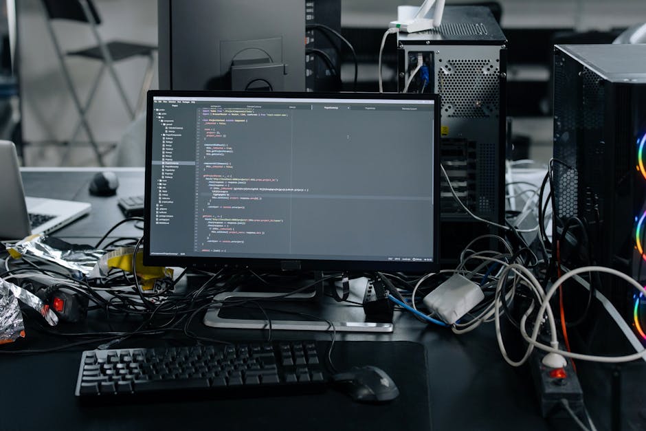 A close-up view of a person's hands operating a laptop on a dark table, with the screen displaying lines of code in a code editor. The background is blurred, highlighting the focus on the laptop and h